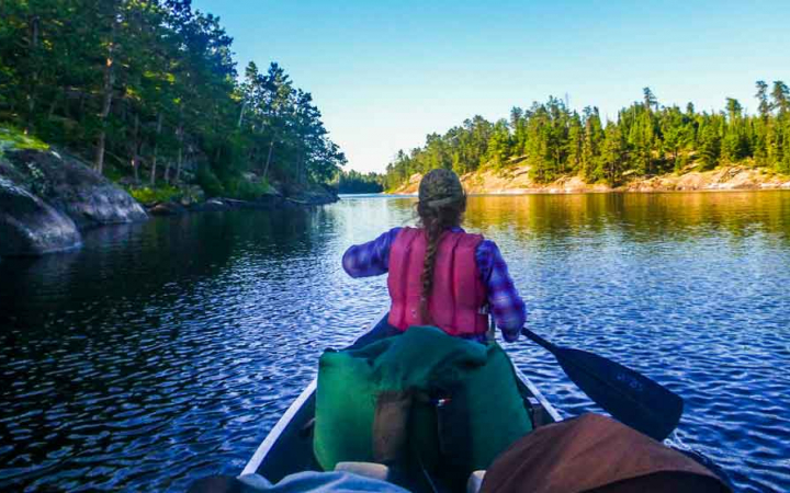 a young person with a long braid sits in the front of a canoe holding a paddle, facing away from the camera. There are evergreen trees lining the shore.